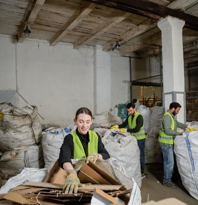 Positive worker in gloves and vest putting cardboard in sack while separating trash near blurred multiethnic colleagues in waste disposal station, garbage sorting and recycling concept